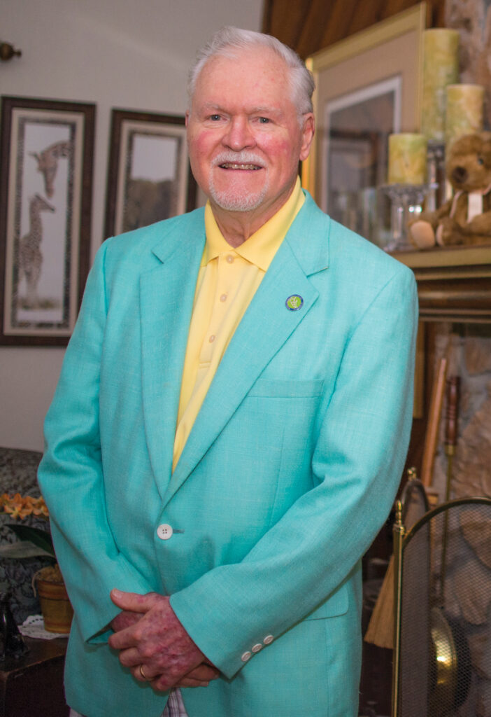 Dr. Whitney at home standing in front of his fireplace with an aqua dinner jacket on.